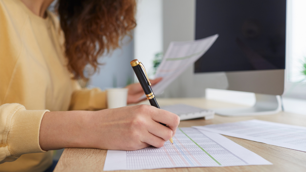 Femme travaillant sur des documents financiers à un bureau, analysant des données avec un stylo à la main, devant un écran d'ordinateur