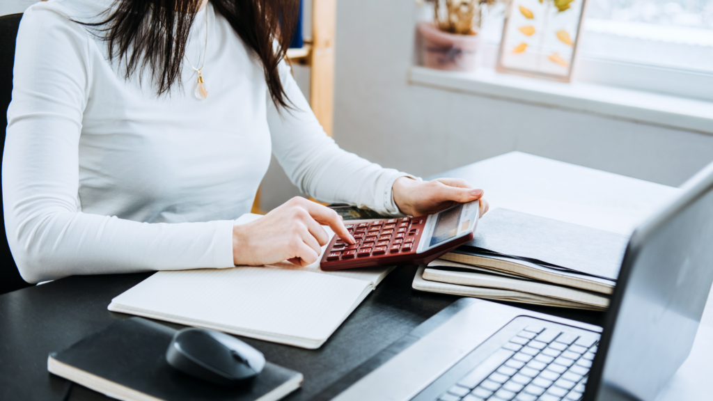 Femme utilisant une calculatrice et un ordinateur portable, travaillant sur des graphiques financiers à un bureau avec une tasse de café à proximité